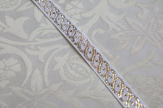 Close-up of white jacquard ribbon with repeating metallic gold scroll and oval design